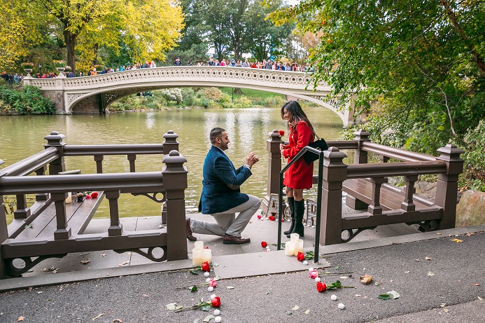 Man down on one knee proposing to girlfriend on decorated landing Bow Bridge