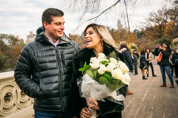 Girlfriend smiles at fiance and holds a dozen white roses after marriage proposal Bow Bridge