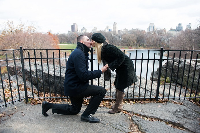Man on one knee kisses fiance at Belvedere Castle with city skyline in background